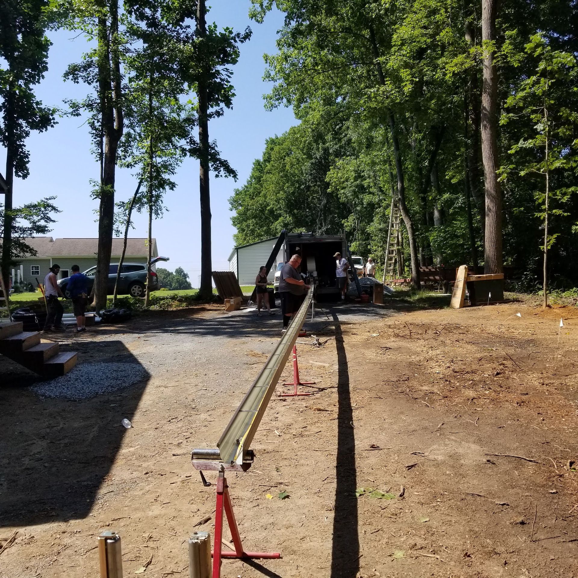 Construction site: crew working with a long metal beam supported by sawhorses. Trees and a vehicle in background.