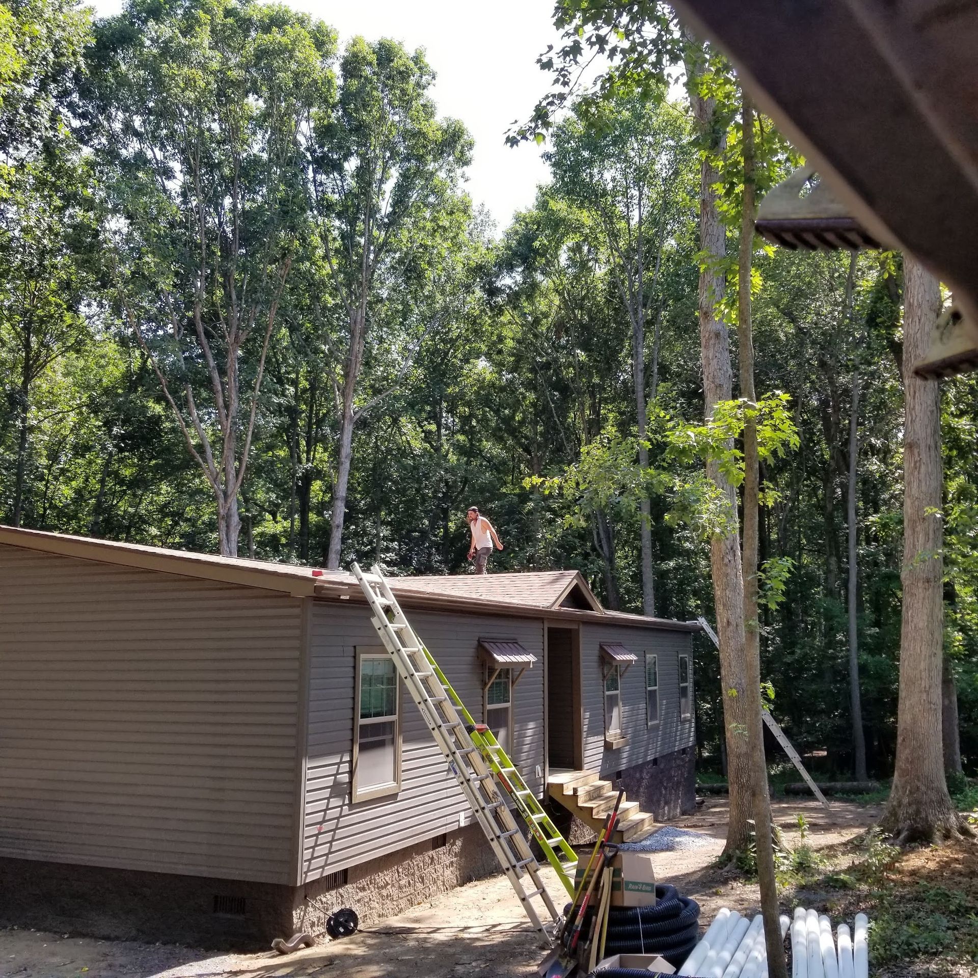 Person on the roof of a house under construction. A ladder is leaning against the side. Trees in the background.