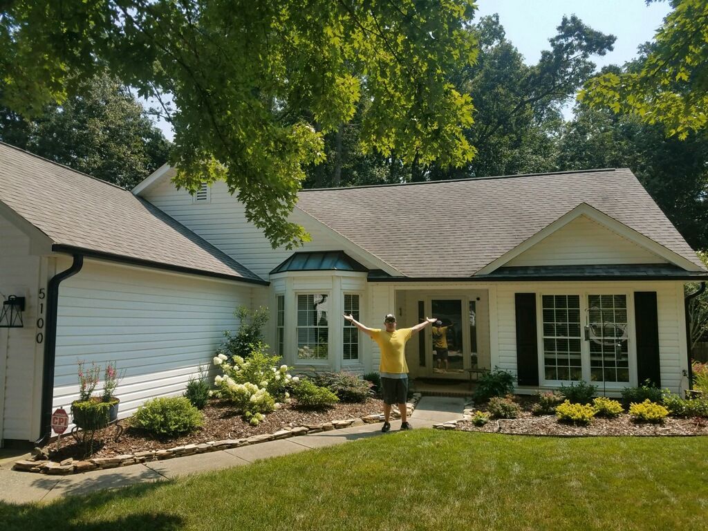 Man with arms outstretched in front of a white house with black shutters, landscaping, and a green lawn.
