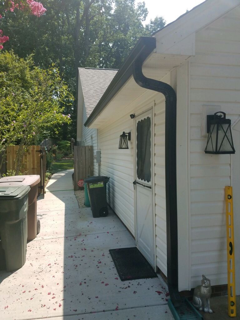 Side of a white building with black gutters and door. Trash cans and a level are visible.