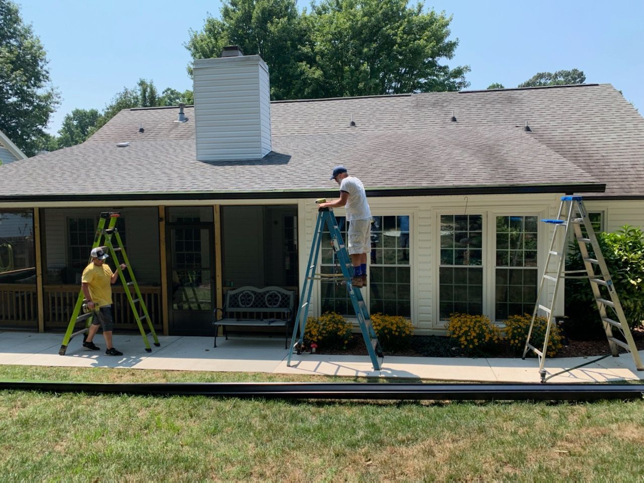 Three workers on ladders installing gutters on a house. Green grass and blue sky are visible.