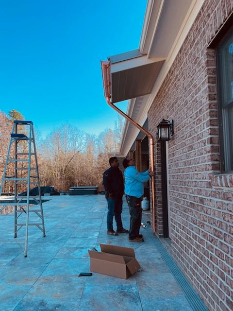 Two workers install copper gutter on a brick house. A ladder, cardboard box, and blue sky visible.