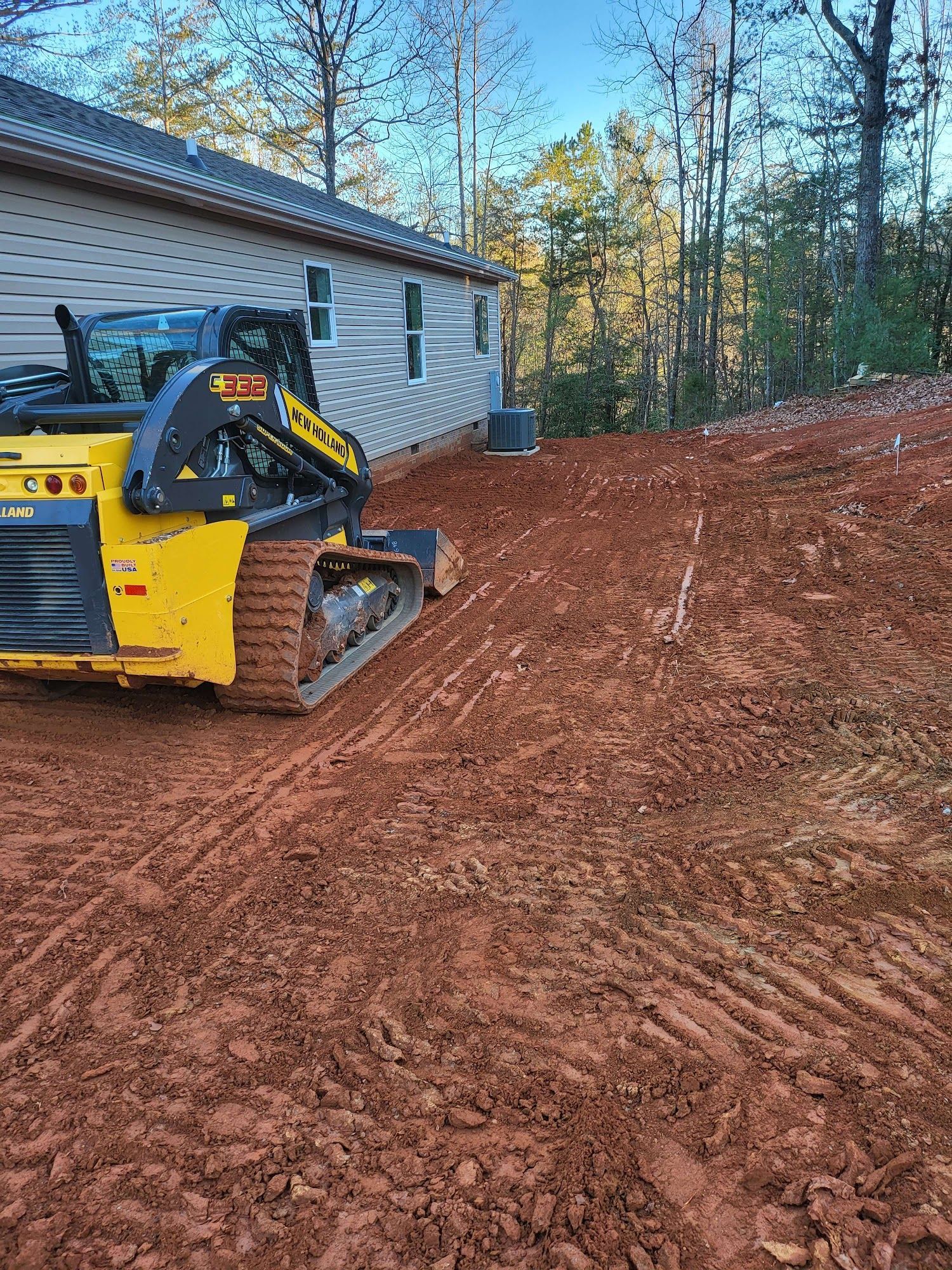 Yellow skid steer on red dirt slope next to a house, with trees in the background.