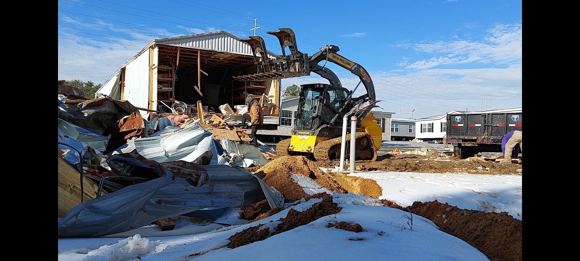 A yellow excavator demolishes a building on a snowy, sunny day. Debris surrounds the machine.