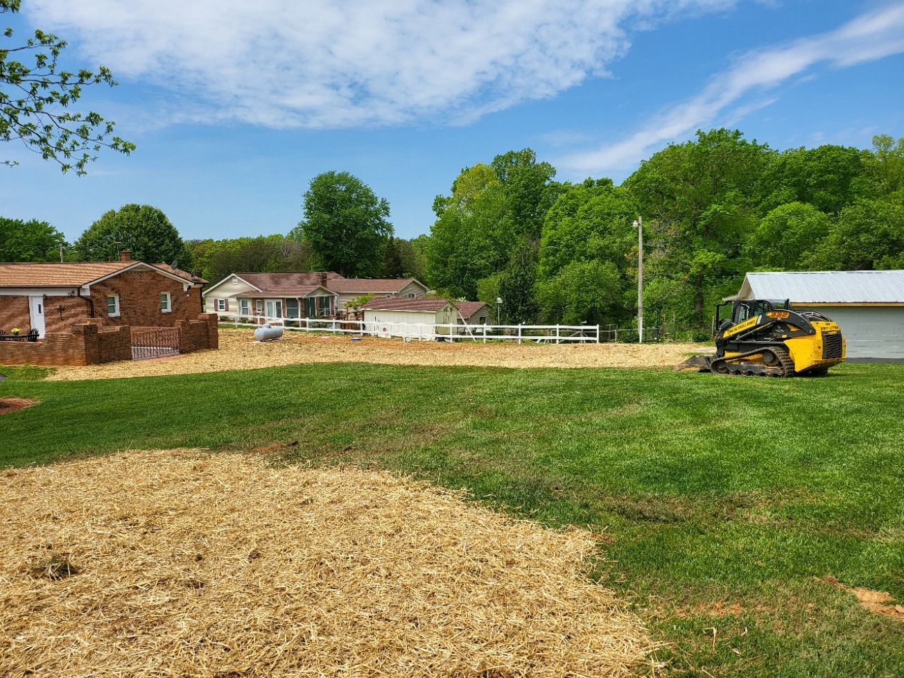 Clearing of land: a yellow bulldozer, house, brown mulch, and green grass against a blue sky.