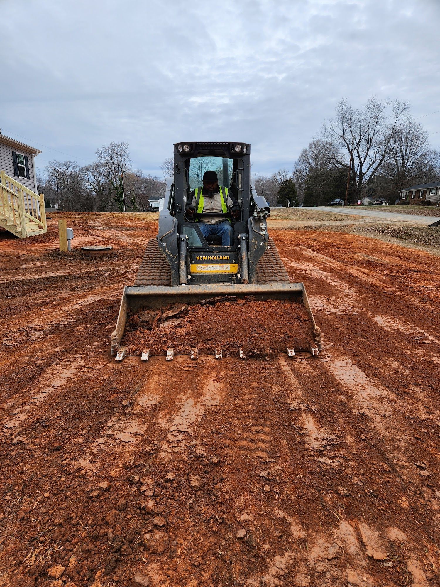 A worker operating a skid steer grading reddish-brown soil in an outdoor construction area.