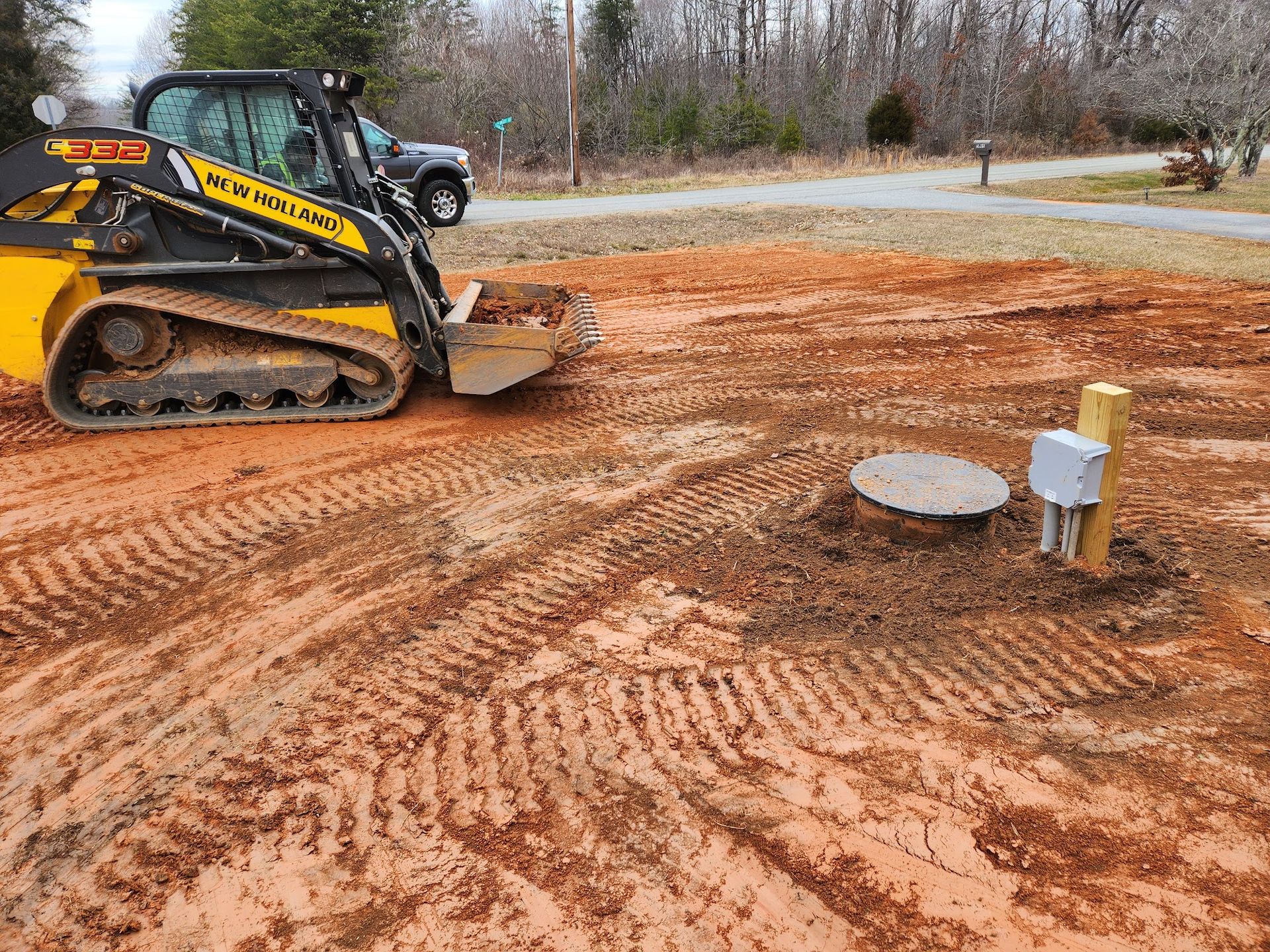 Skid steer loader next to an open septic tank in a dirt yard. Electrical box on a wooden post.