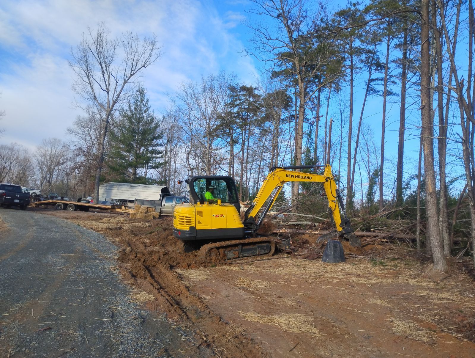 Yellow excavator on muddy ground, clearing debris near trees and road under a blue sky.