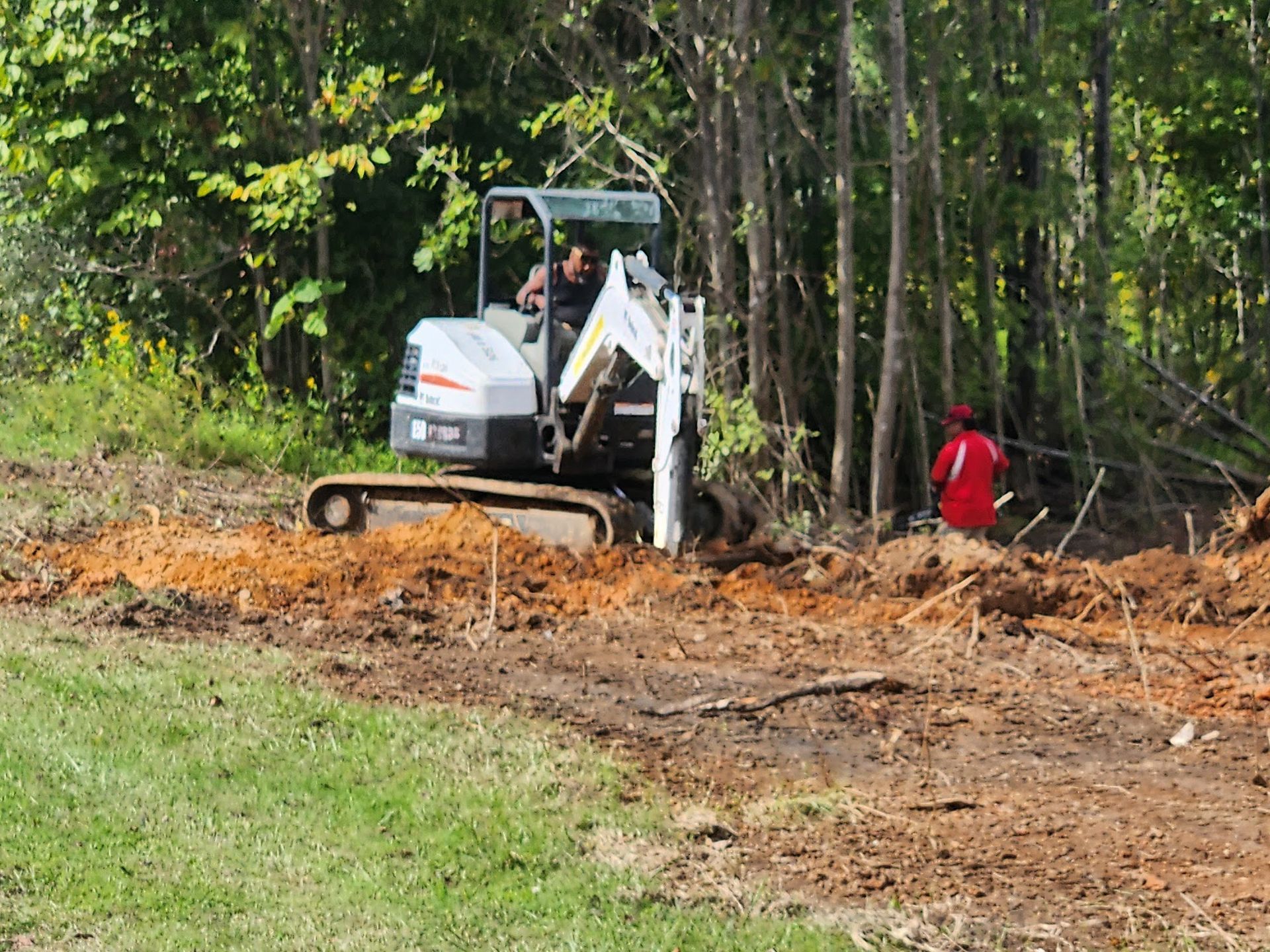 A small excavator digs in dirt near a treeline; a person in a red shirt stands nearby.