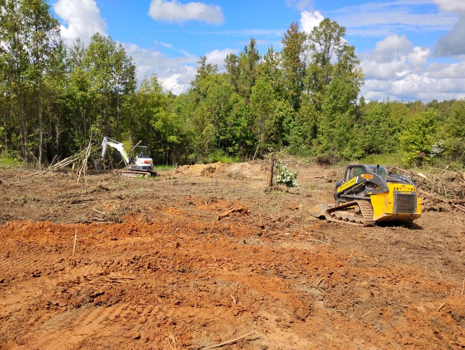 Two construction vehicles on a cleared dirt lot in front of a line of trees under a blue sky.