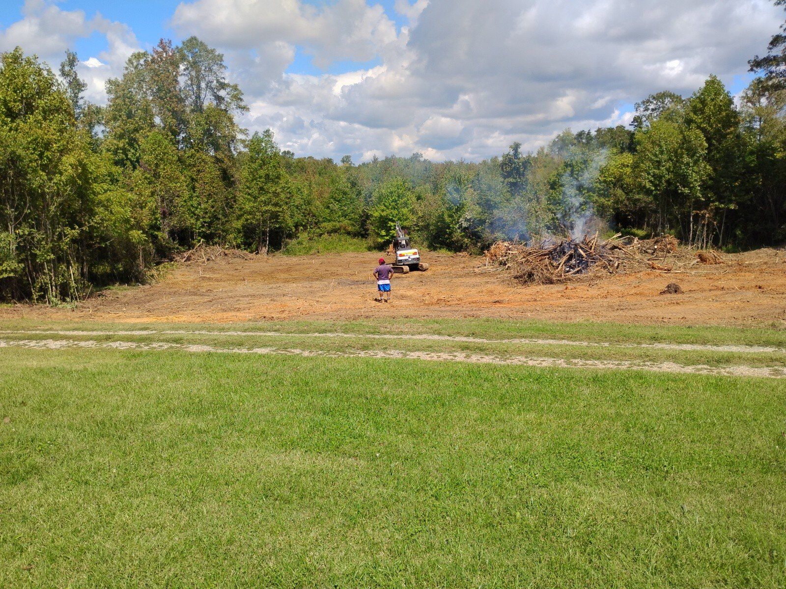 A person walks toward a clearing where brush is burned. Trees and a white truck are also visible under a cloudy sky.