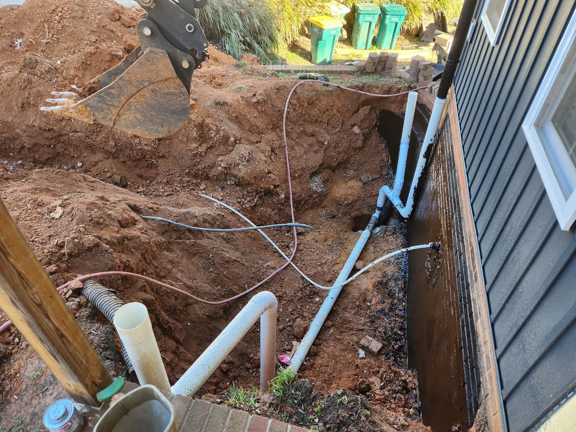Excavation site with plumbing pipes and excavator bucket near a house wall, exposed soil, and trash cans.