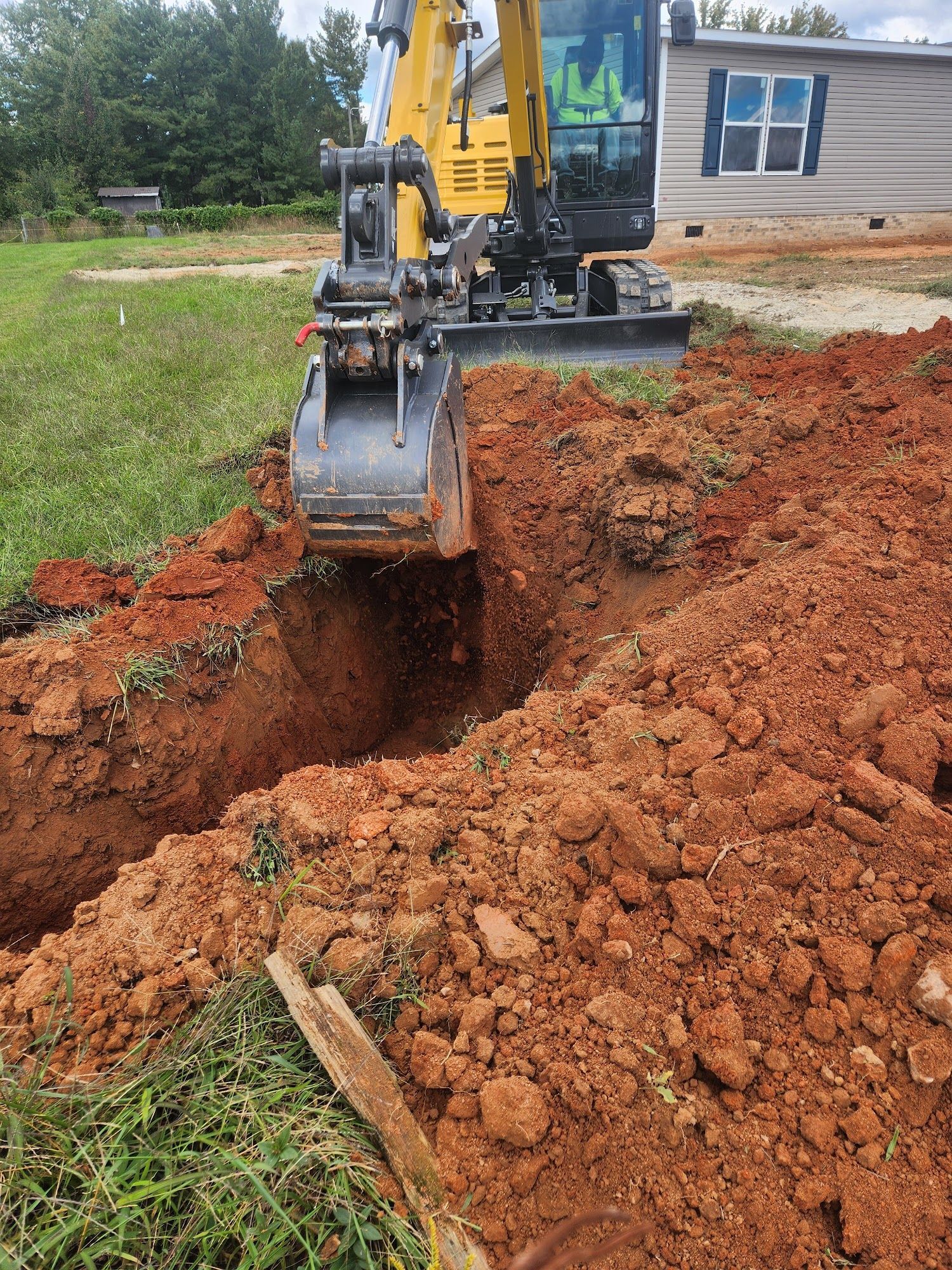 Yellow excavator digging a trench in red soil near a house.