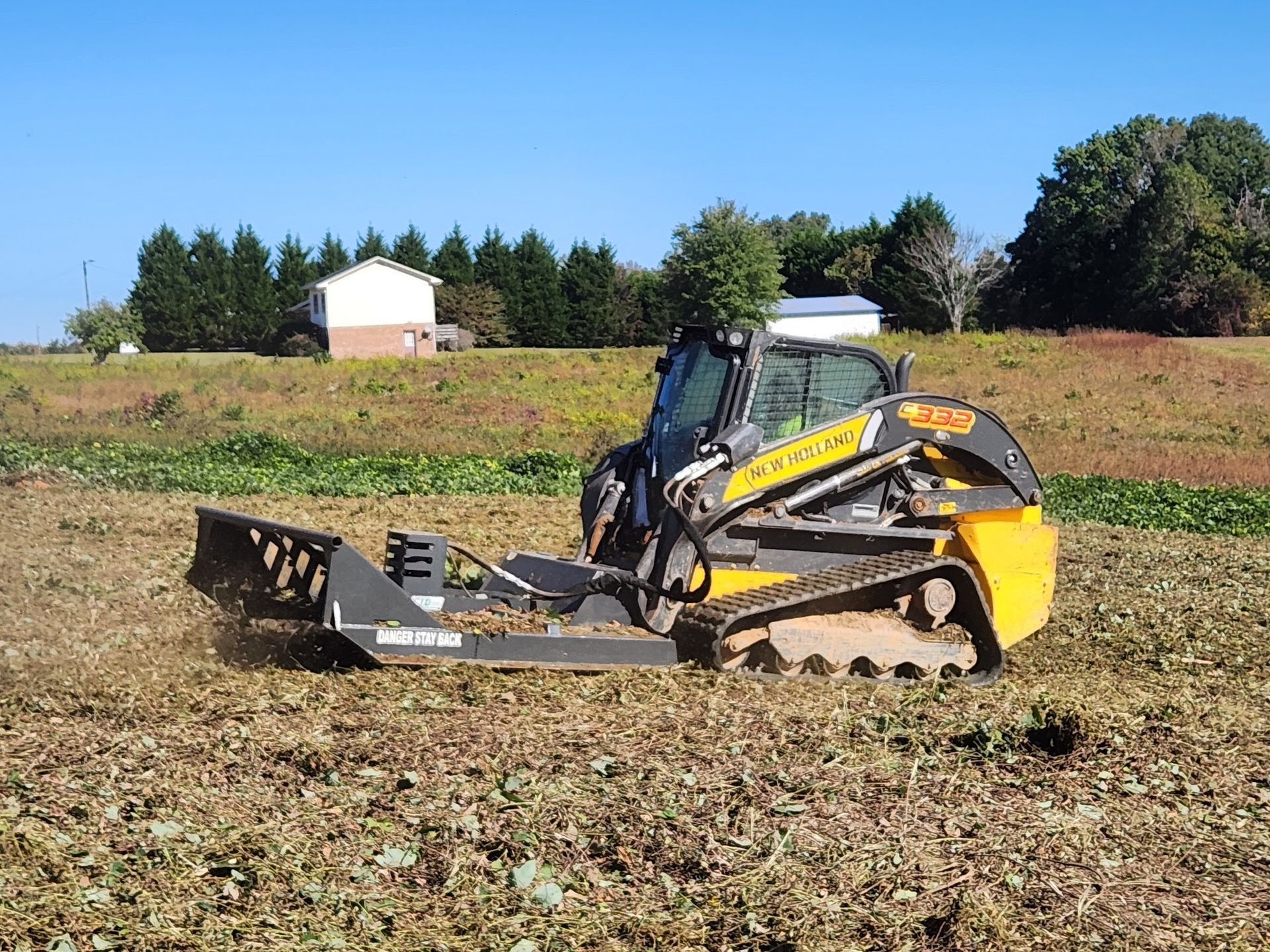 Skid steer with brush cutter mulching vegetation in a field on a sunny day.