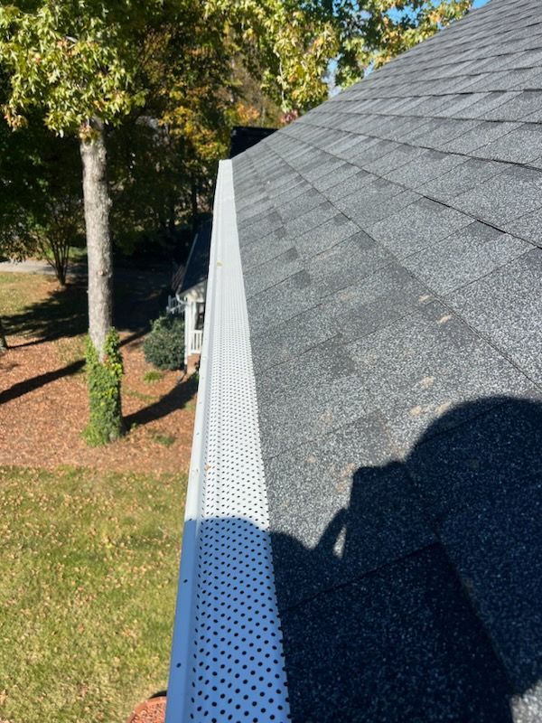 Gutter with a leaf guard on a dark shingle roof, green lawn, and trees.