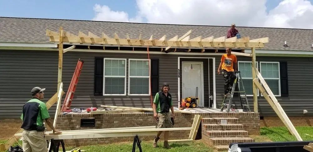 Workers building a porch awning on a house. Wooden beams and supports are in place, with multiple workers involved.