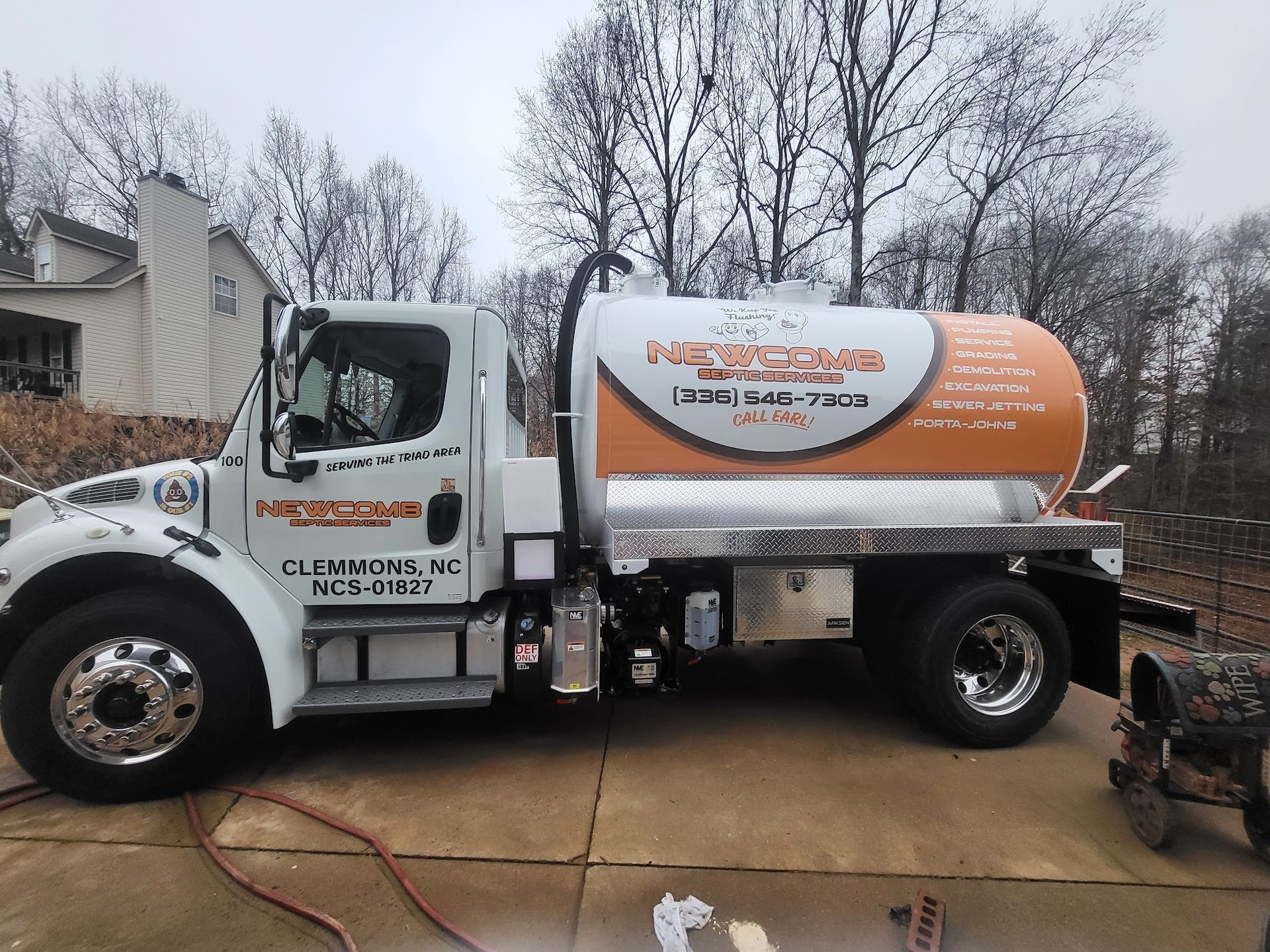 White and orange septic service truck parked on a concrete surface with a house in the background.