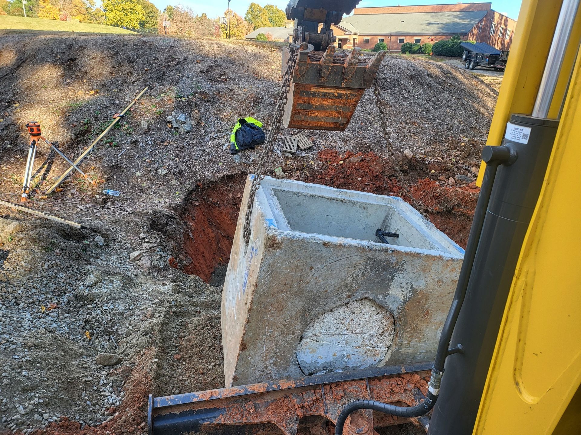 Yellow excavator digging a trench in red dirt, house in background.