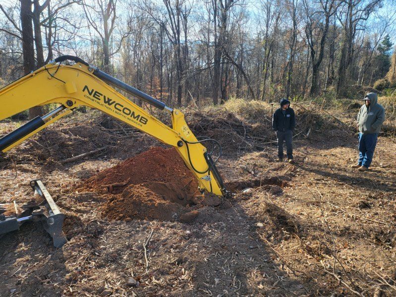 A small excavator digs in dirt near a treeline; a person in a red shirt stands nearby.