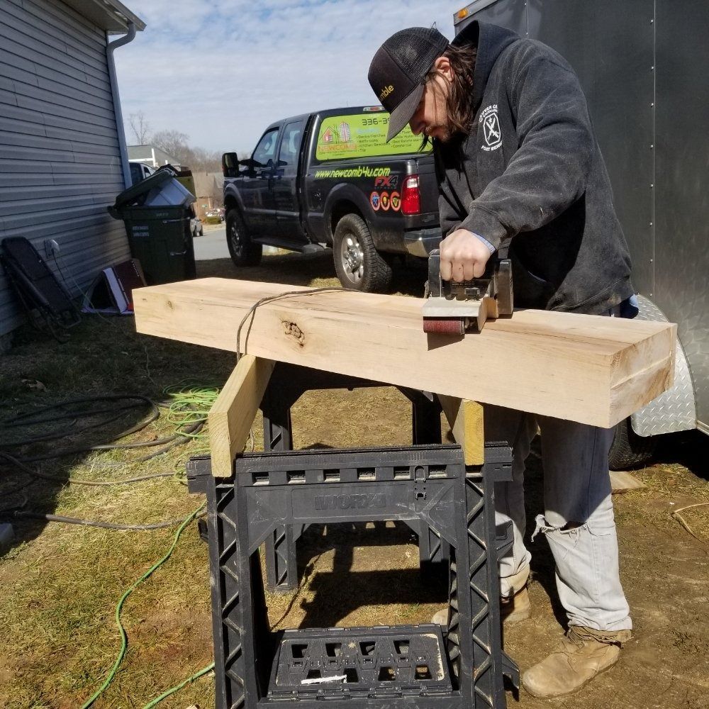 A person using a power planer on a wooden beam, outdoors, with a truck and trailer in the background.