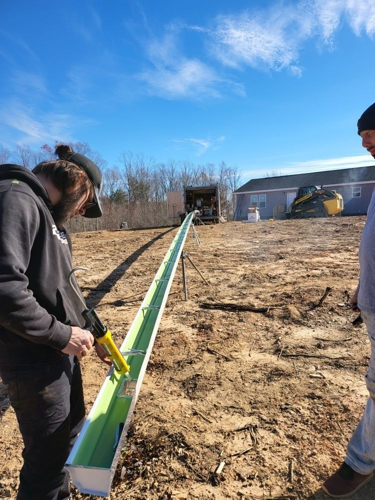 Two men work on a long, green gutter in a field, one using a caulking gun. Sunny day.