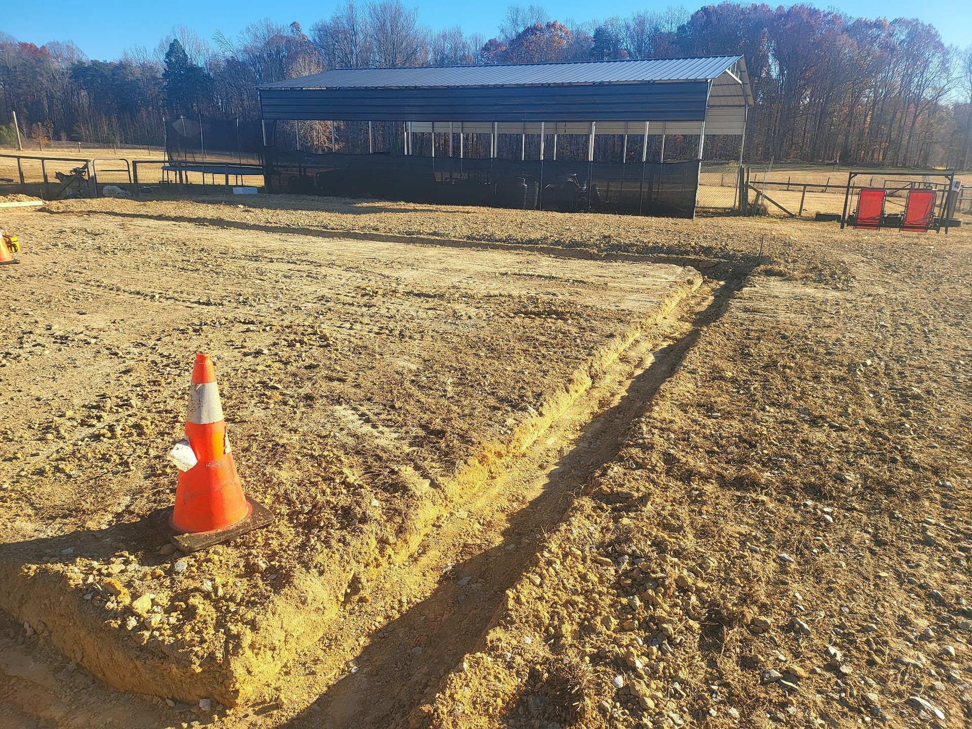 A trench dug in a dirt field with a barn in the background and a traffic cone.