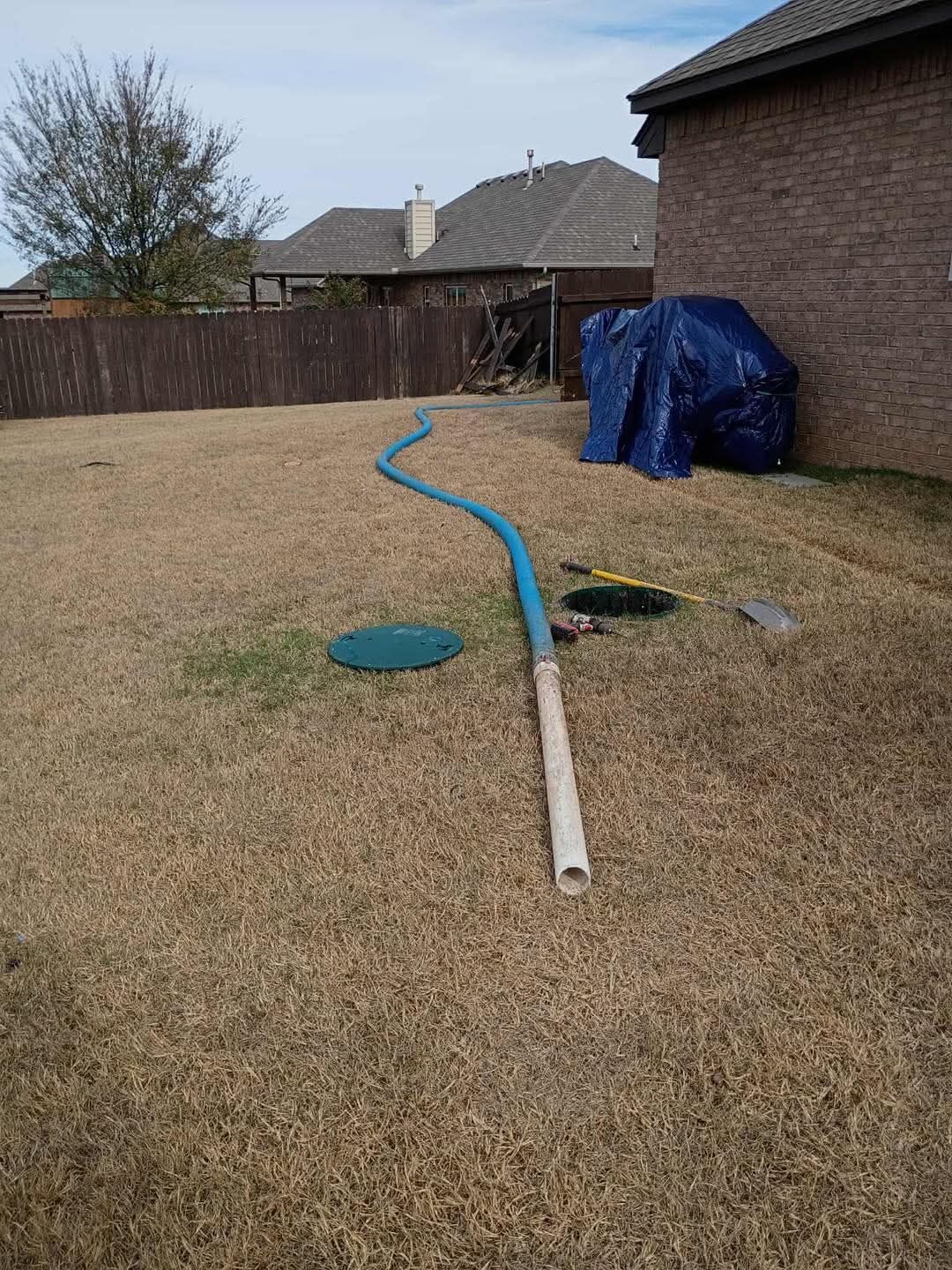 A backyard with a blue hose, a septic tank lid, and dry grass. A tarp covers something by the brick house.