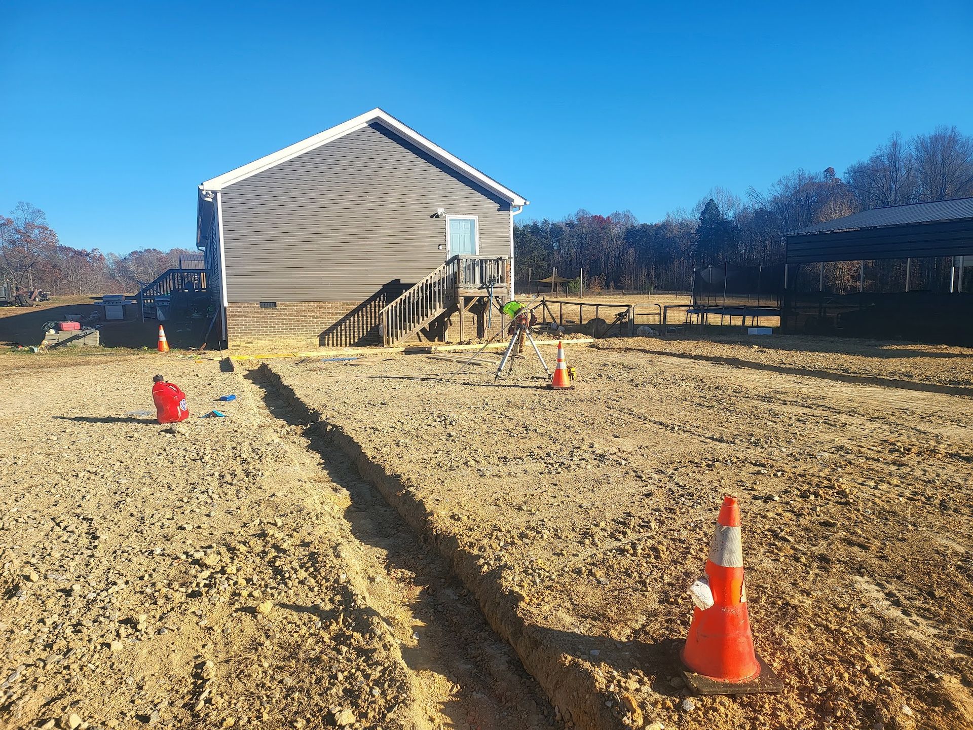 A trench is dug in front of a house, marked with orange cones and containers, clear blue sky.