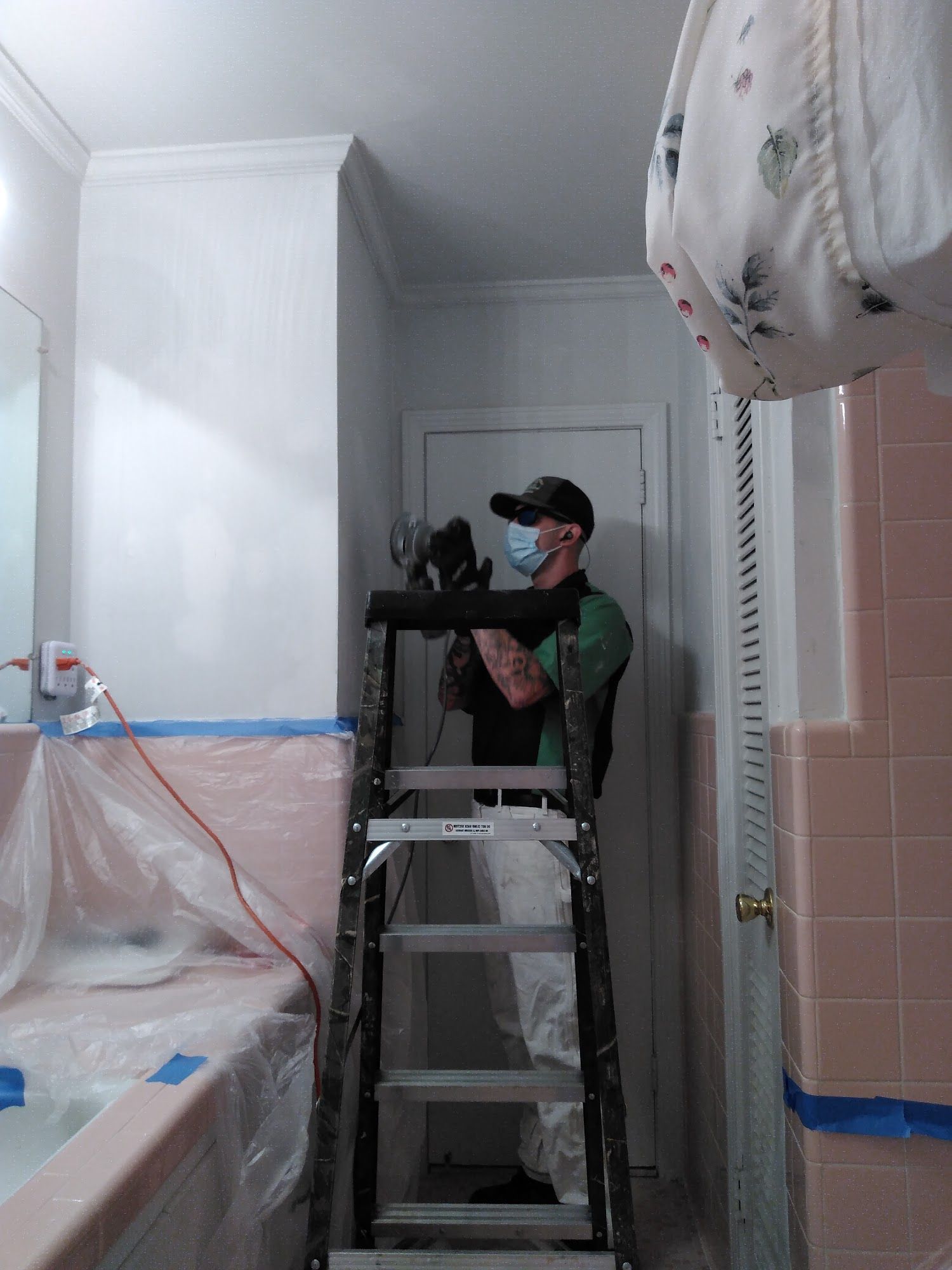 Man on ladder painting bathroom walls, wearing mask and protective gear. Pink bathtub and tiles are covered in plastic.