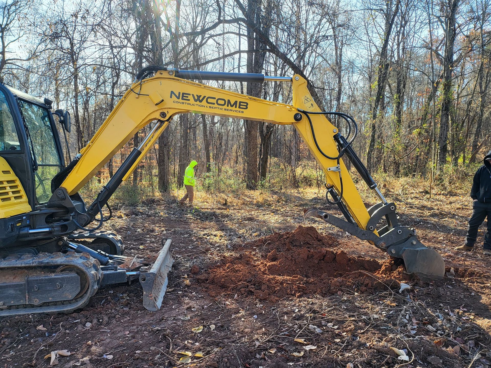 Yellow excavator digging in a wooded area with two people standing nearby.