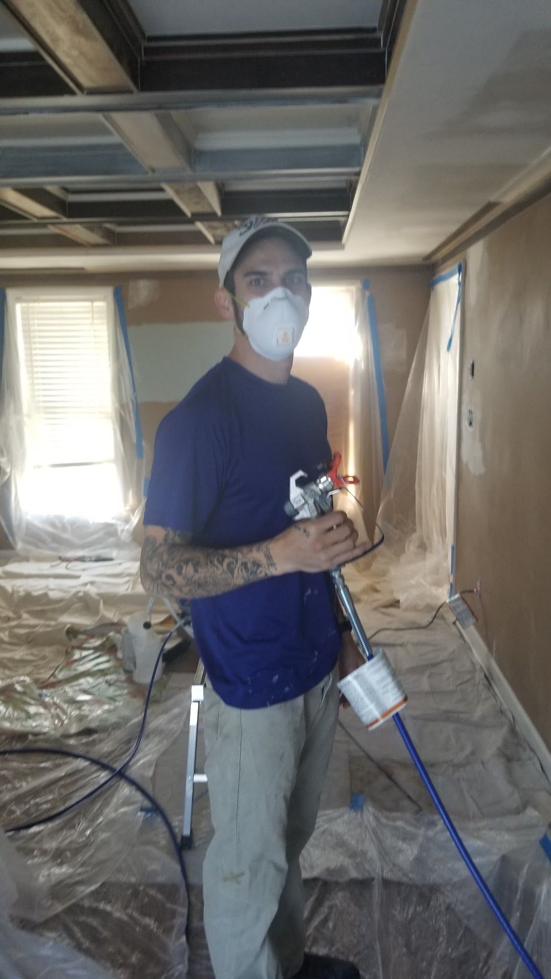 Person in a mask holding a paint sprayer, standing in a room covered in plastic during renovation.