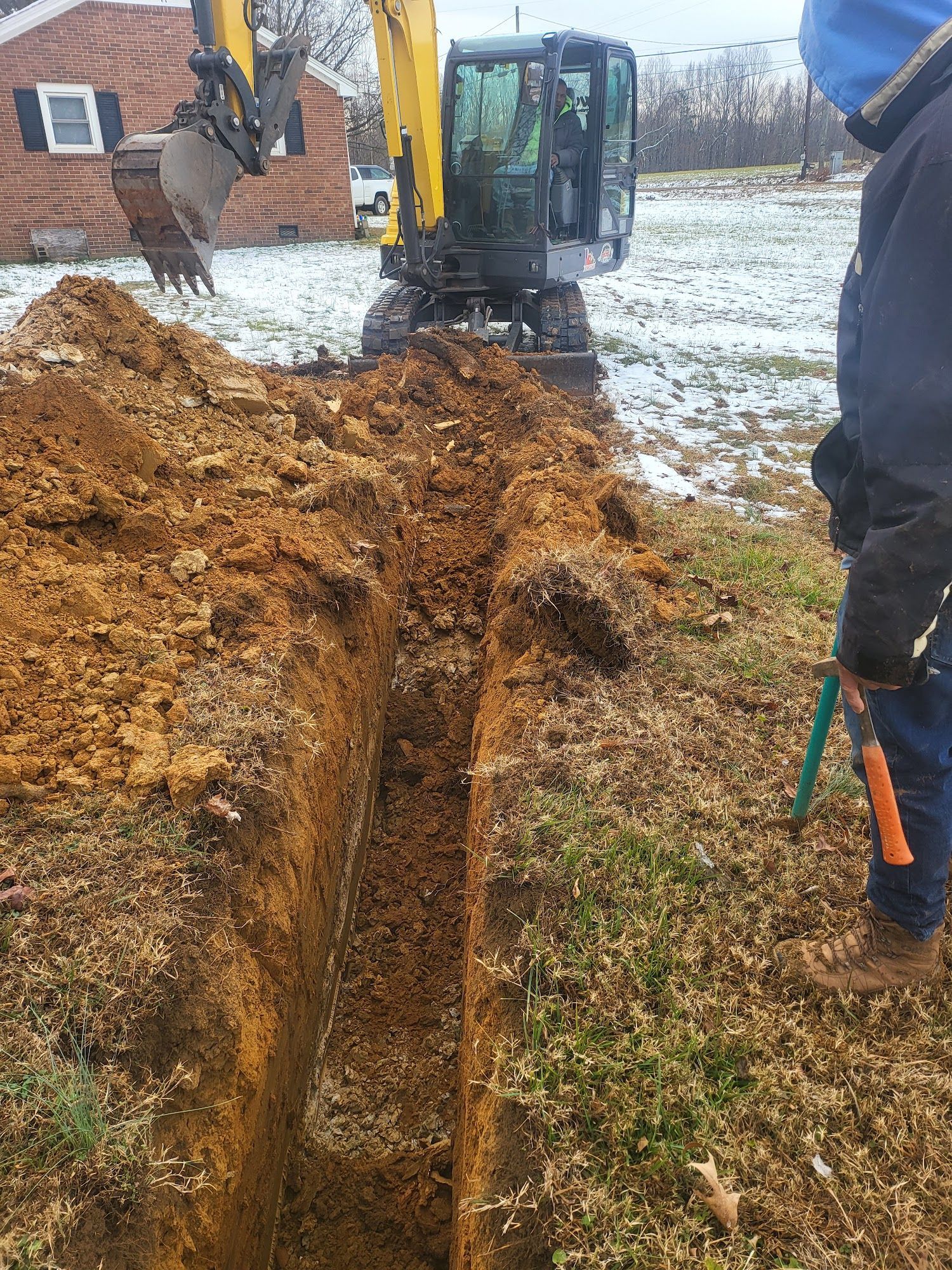 A trench being dug by an excavator on a grassy area, with a person holding a tool nearby.