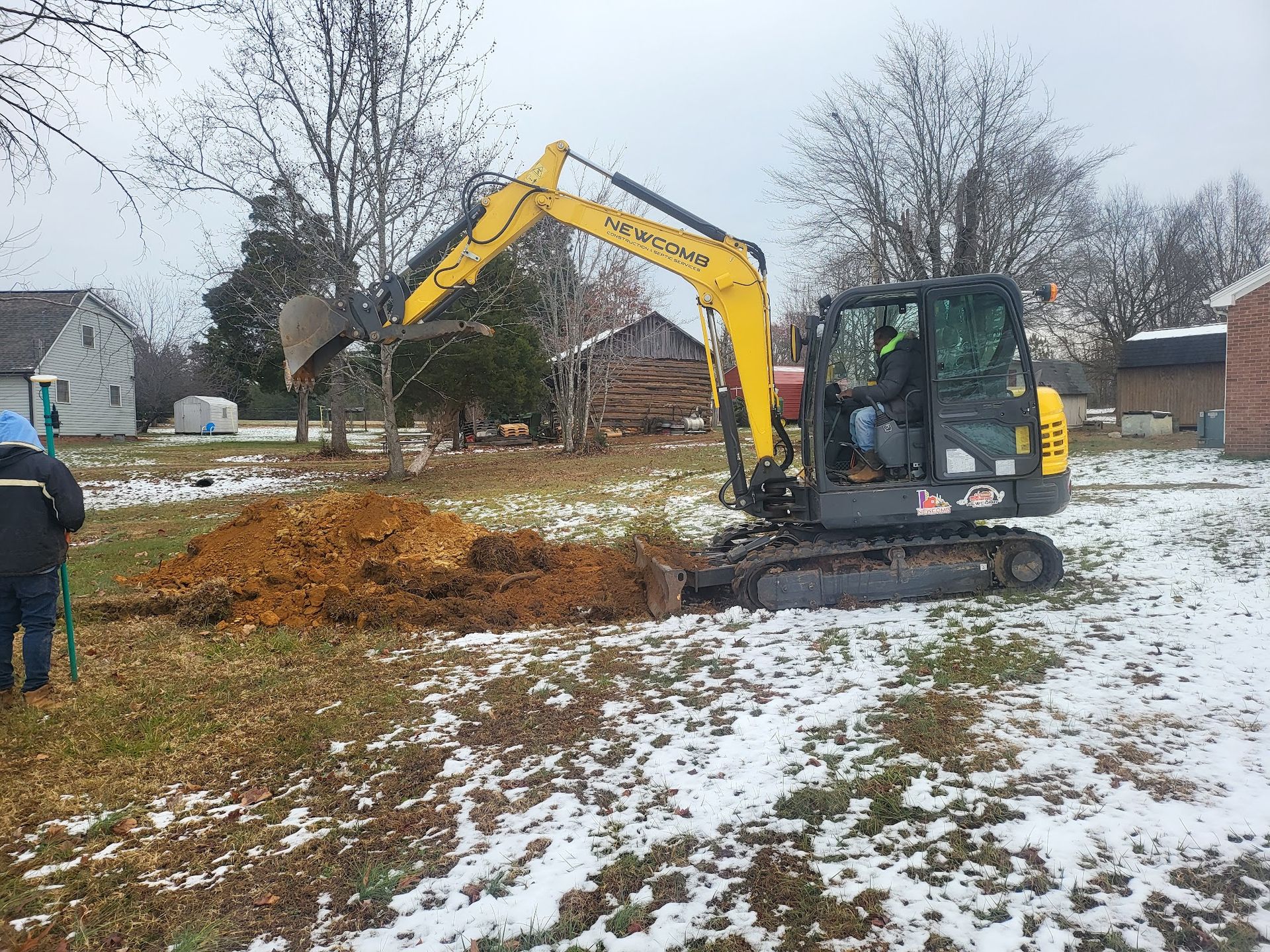 A yellow excavator digs in a snowy yard, with a person operating it. Another person watches nearby.