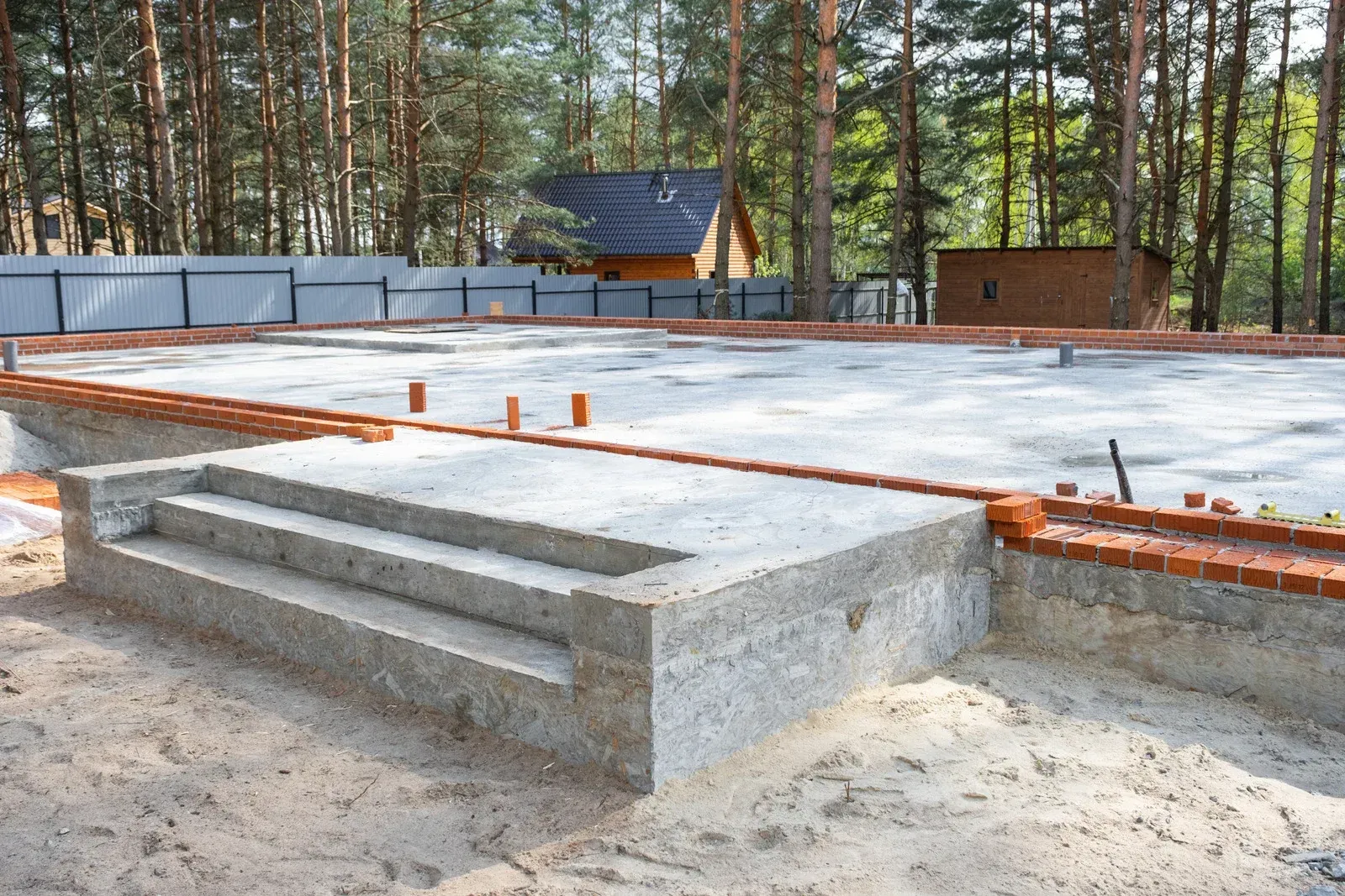 Construction site with concrete foundation and steps, surrounded by sand and brick, in a wooded area.