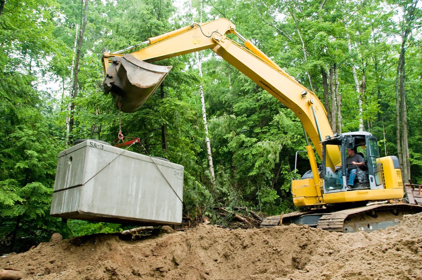 Yellow excavator lifting a large concrete septic tank into an earthen hole, in a wooded area.