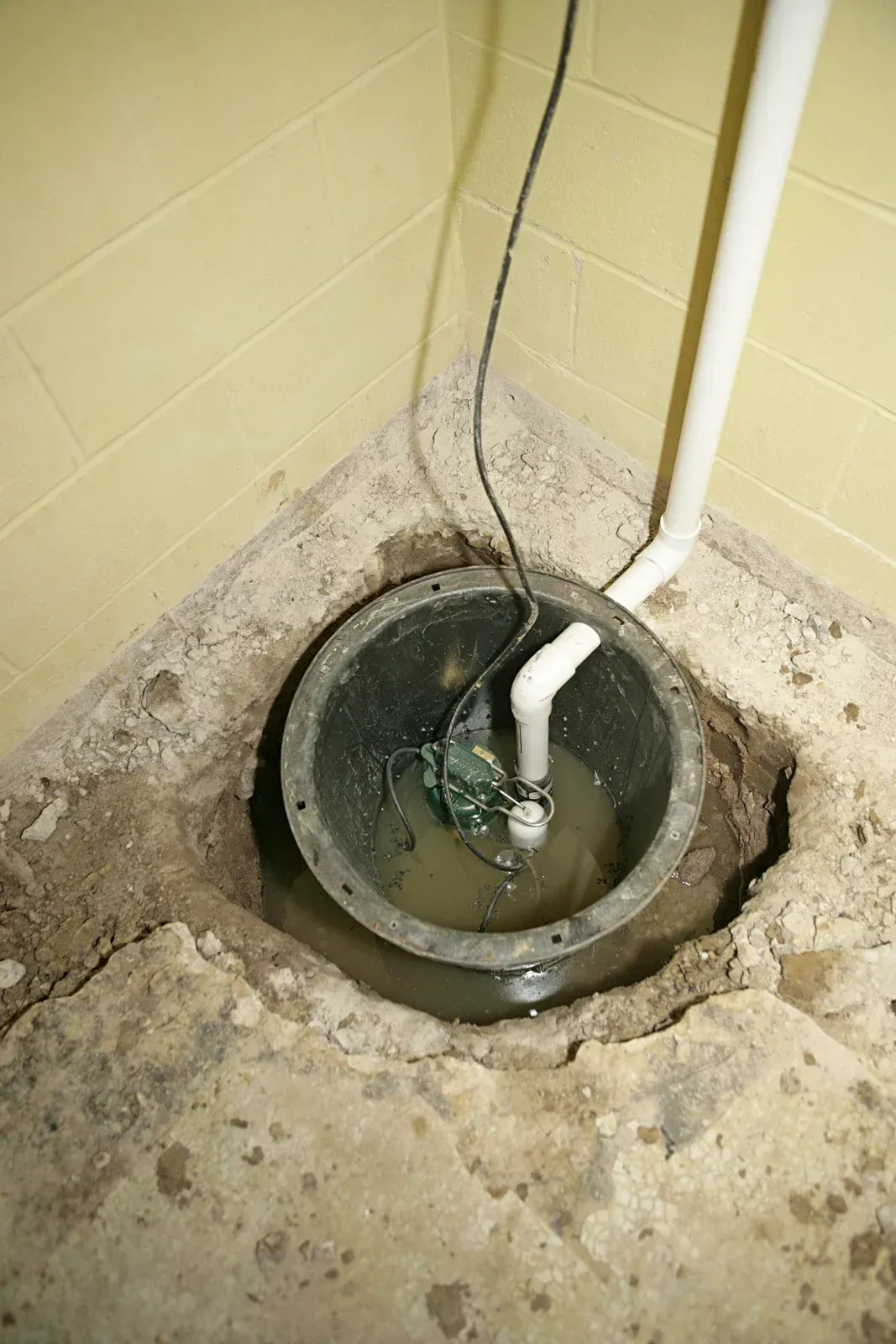 Sump pump in a concrete pit. Black pump with a white discharge pipe in standing water, in a basement corner.