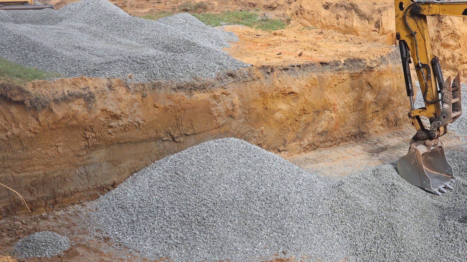 Yellow excavator dumping gravel onto a gravel surface. Dirt mound in the background.