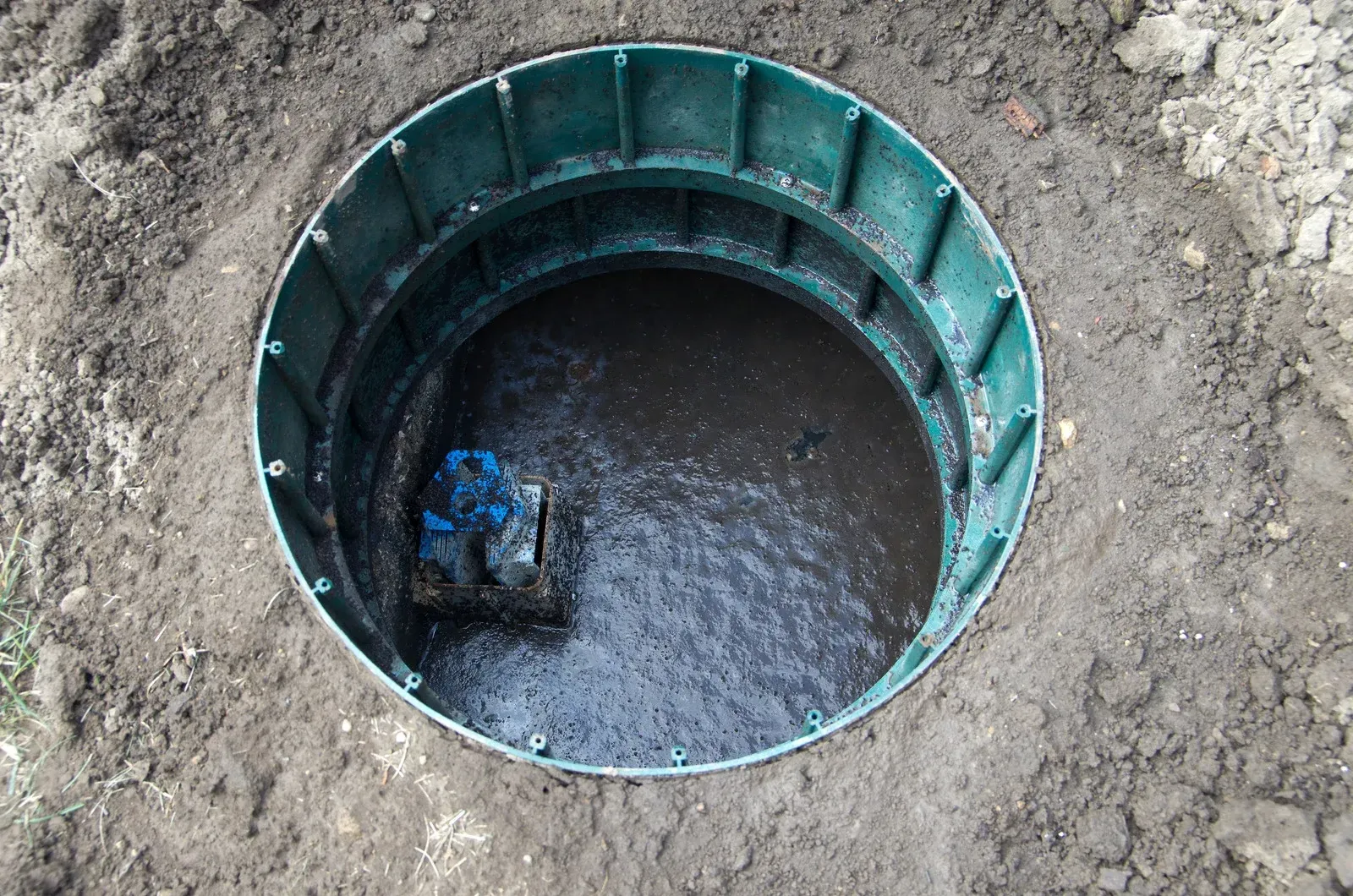 Open green septic tank surrounded by dirt, filled with dark liquid, visible pipe.