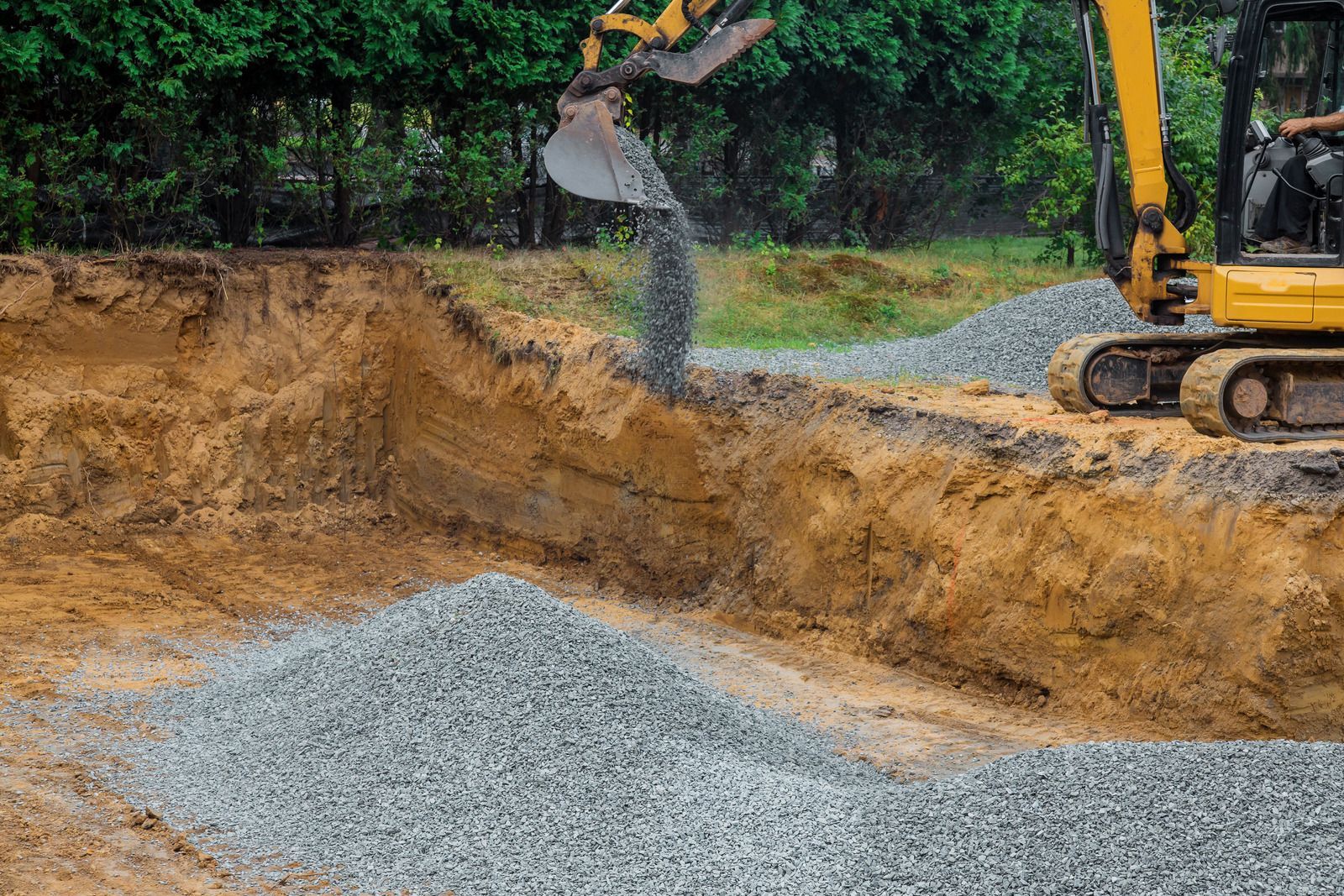 Yellow excavator pouring gravel into an excavated foundation. Earth tones and greenery in the background.