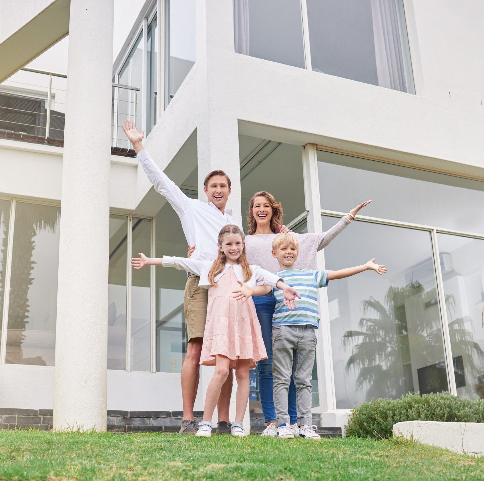 Family of four standing in front of a white house with arms outstretched, smiling.