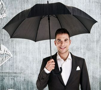 Man in suit smiles, holding a black umbrella open against a gray wall.