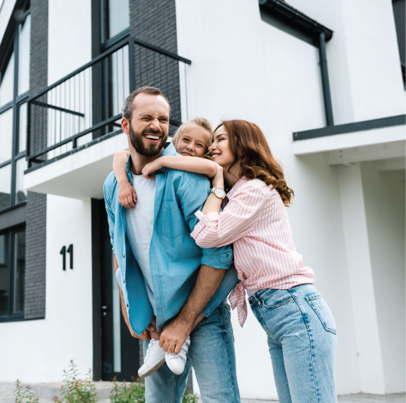 Family in front of a house: Father giving daughter a piggyback ride, mother hugs them, all smiling.