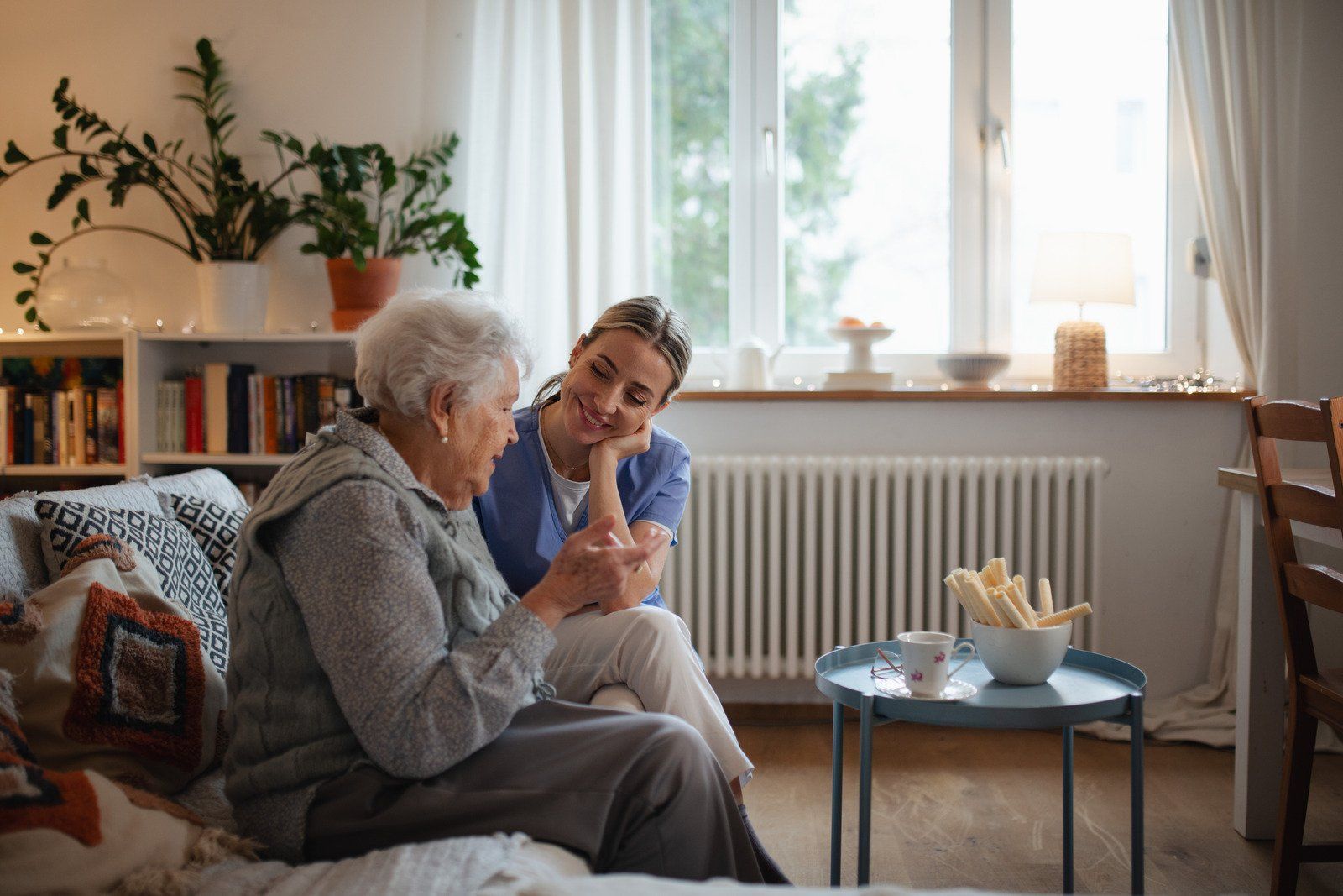 A caregiver in blue scrubs sits on a couch, holding hands with an older person in a bright living room.