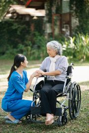 A caregiver in blue scrubs kneels to hold the hands of a smiling person seated in a wheelchair outdoors.
