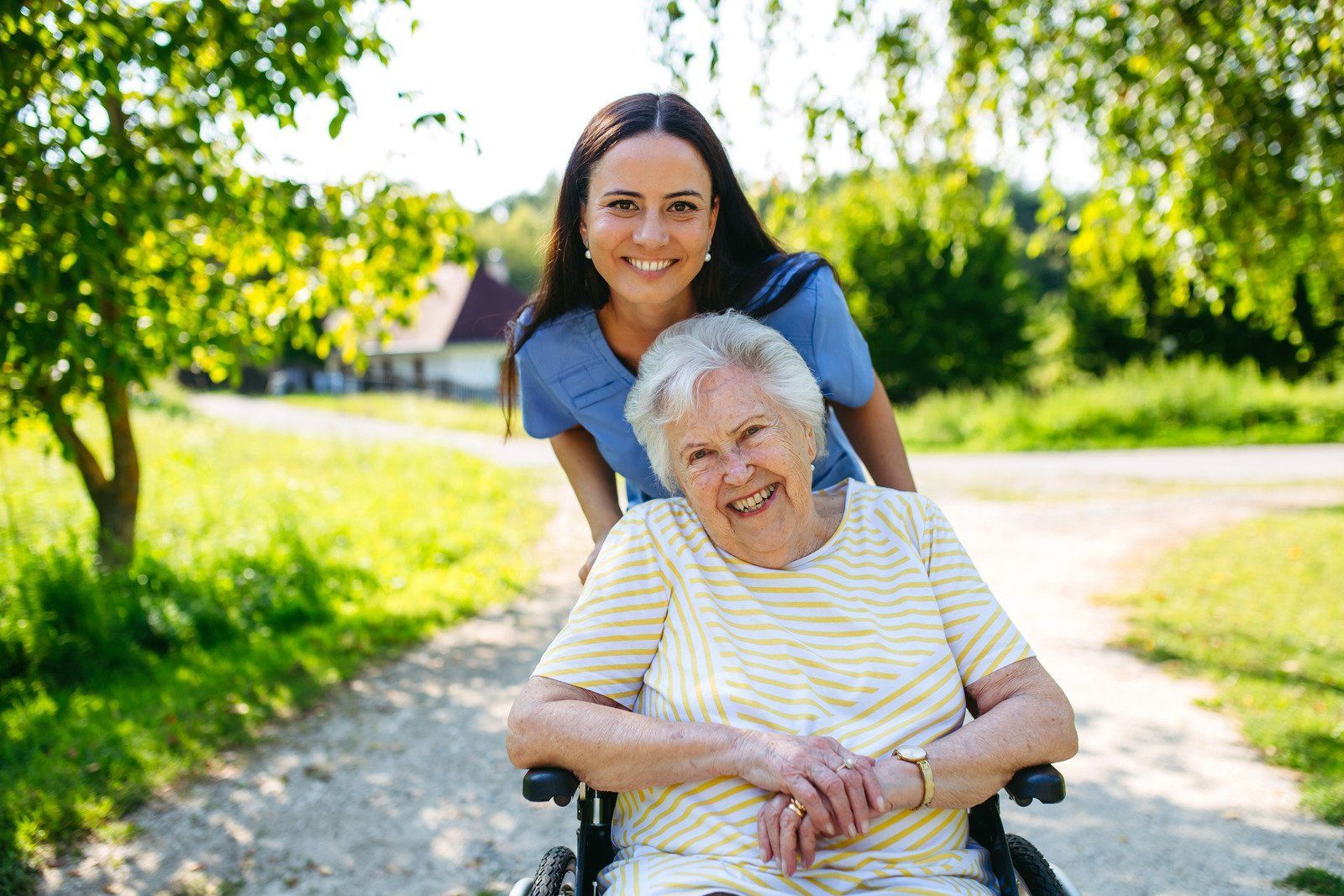 A person in a wheelchair smiles outdoors with a caregiver standing behind them on a sunny, tree-lined path.
