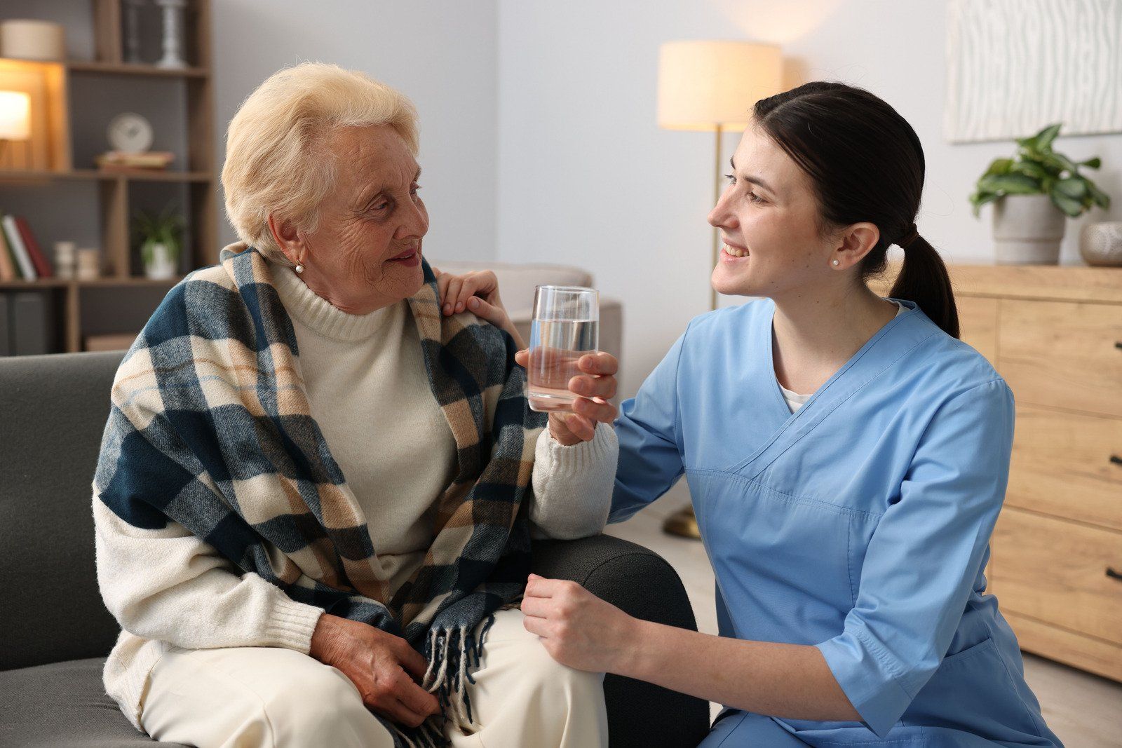 A healthcare worker in blue scrubs offering a glass of water to an older person wearing a shawl in a home setting.