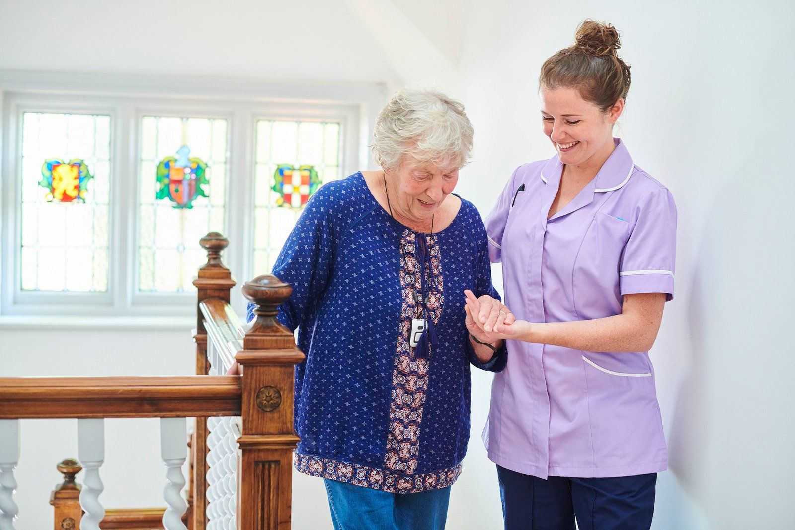 A caregiver in a lavender uniform provides support to an older person walking down a staircase by holding their hand.