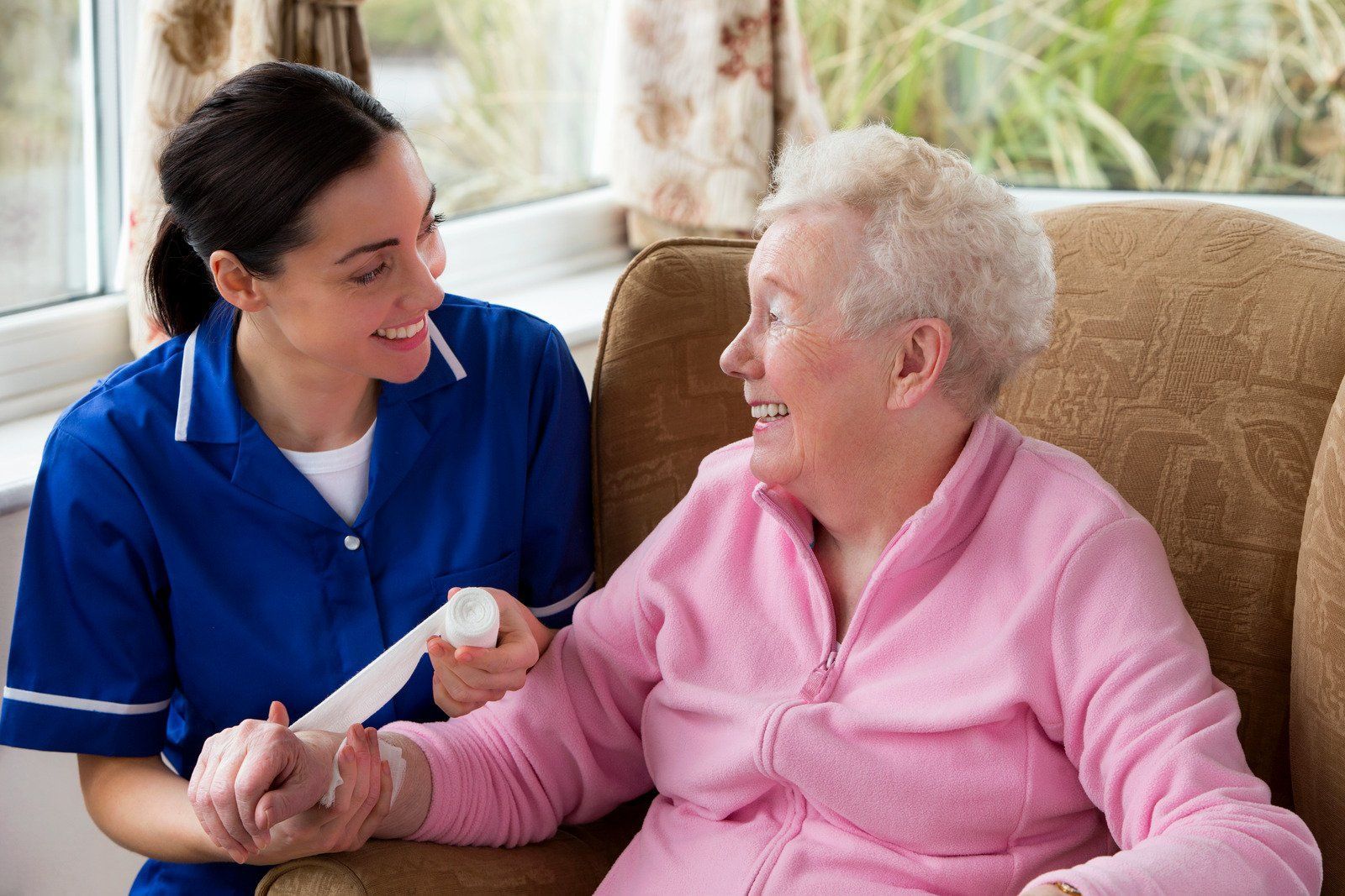 A caregiver in a blue uniform bandages the arm of a person wearing a pink sweater while they smile at each other.
