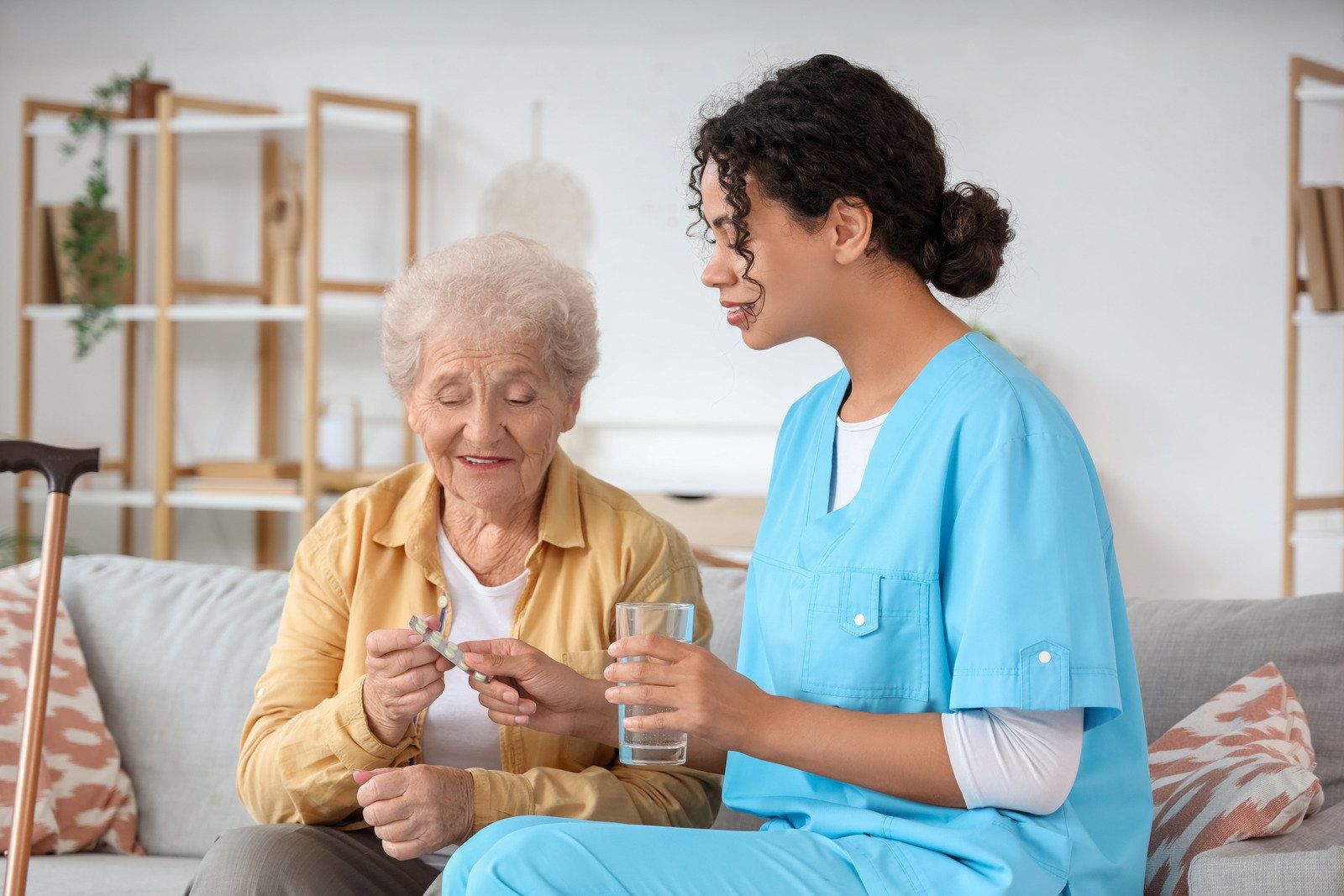 A caregiver in blue scrubs hands a glass of water to an older person holding medication on a sofa.