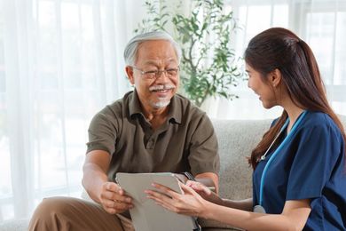 A healthcare provider in blue scrubs shows a tablet to a smiling person seated on a sofa.